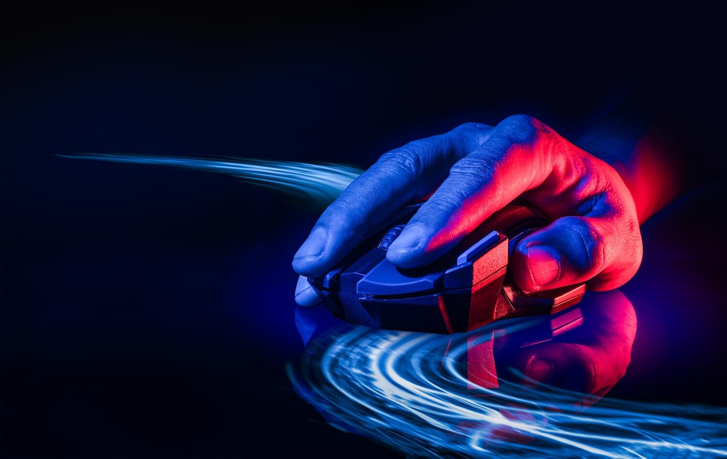 Our Story A hand grips a gaming mouse on a reflective surface, illuminated by blue and red light. Dynamic blue streaks of light trail from the mouse, adding a sense of speed and motion against a dark background.