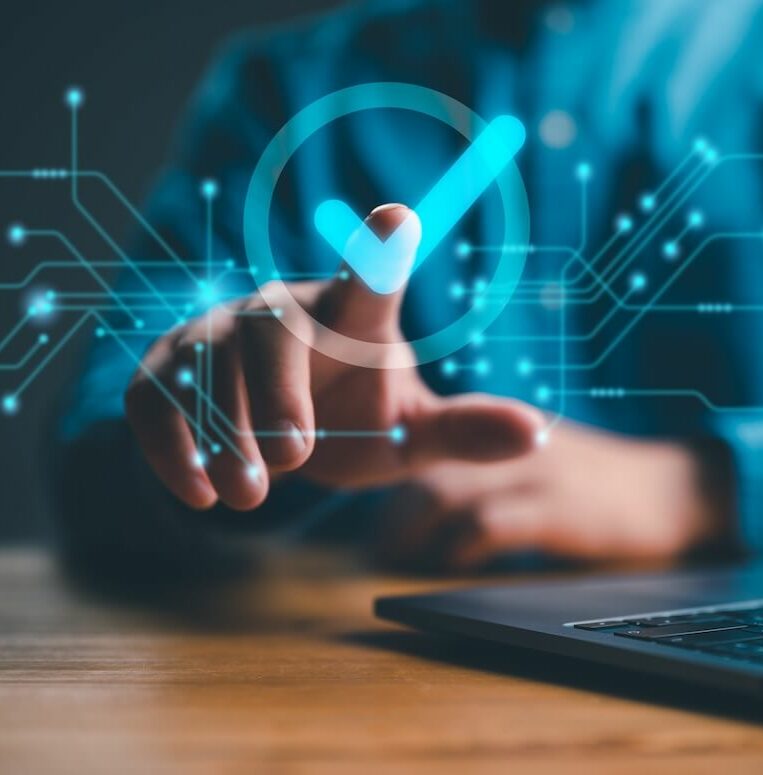 A person is pointing at a glowing checkmark hologram in front of a laptop. The checkmark is surrounded by digital circuit lines, symbolizing technology and approval. The scene conveys a sense of modernity and digital interaction.