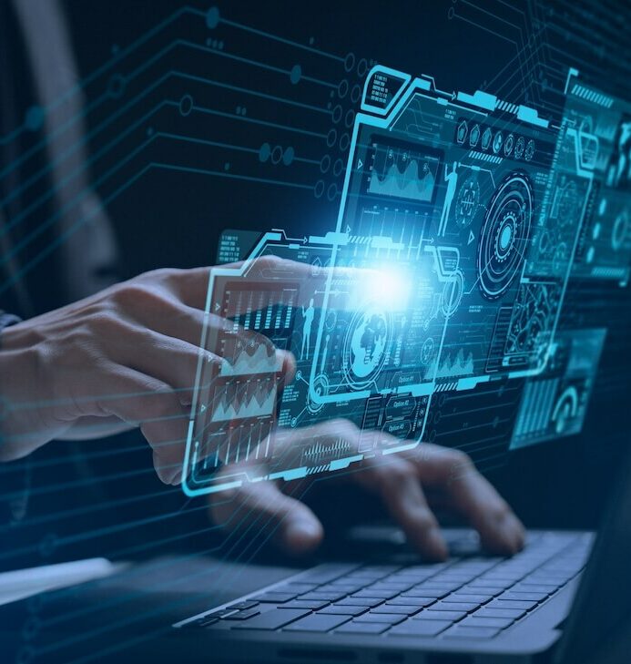 Hands typing on a laptop keyboard with glowing blue digital holograms of various technical interfaces and diagrams projected above the keys, symbolizing advanced technology and innovation.