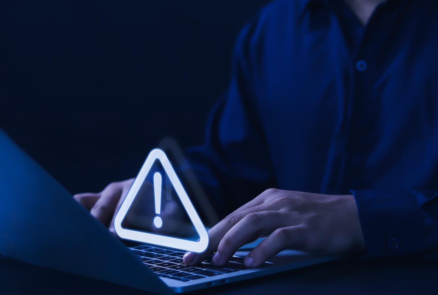 Tired of endless technical issues? Person typing on a laptop in a dimly lit environment. A glowing triangular warning symbol with an exclamation mark hovers above the keyboard, indicating caution or alert. The person is wearing a dark shirt.