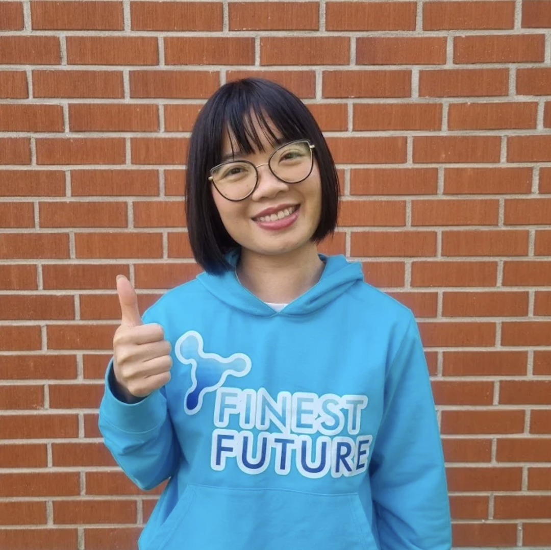 Bambi Dang A person with short black hair and glasses smiles and gives a thumbs up. They are wearing a bright blue hoodie that says "FINEST FUTURE," standing in front of a brick wall.