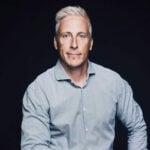 A man with short, light blond hair and light eyes wears a light blue button-up shirt and poses against a plain dark background, looking confidently at the camera.