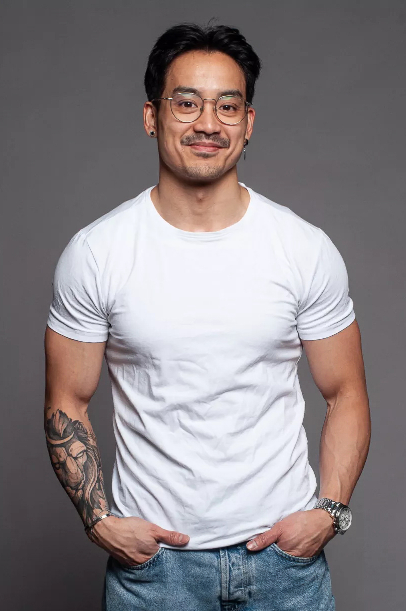 Duy A man with short dark hair, glasses, and a tattoo on his left forearm smiles at the camera. He is wearing a white t-shirt and blue jeans, standing against a plain grey background.