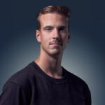 A young man with short, light brown hair and light facial hair wearing a black crew-neck shirt, posing against a gradient dark blue background.
