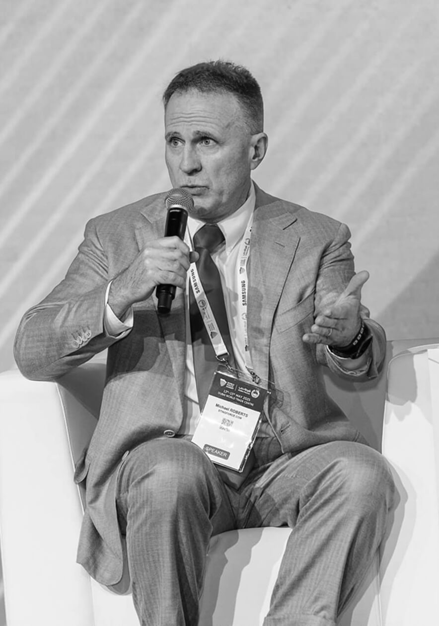 Michael Roberts A man in a suit sits on a white chair, speaking into a microphone. He gestures with his left hand, wearing a conference badge around his neck. The image is in black and white.
